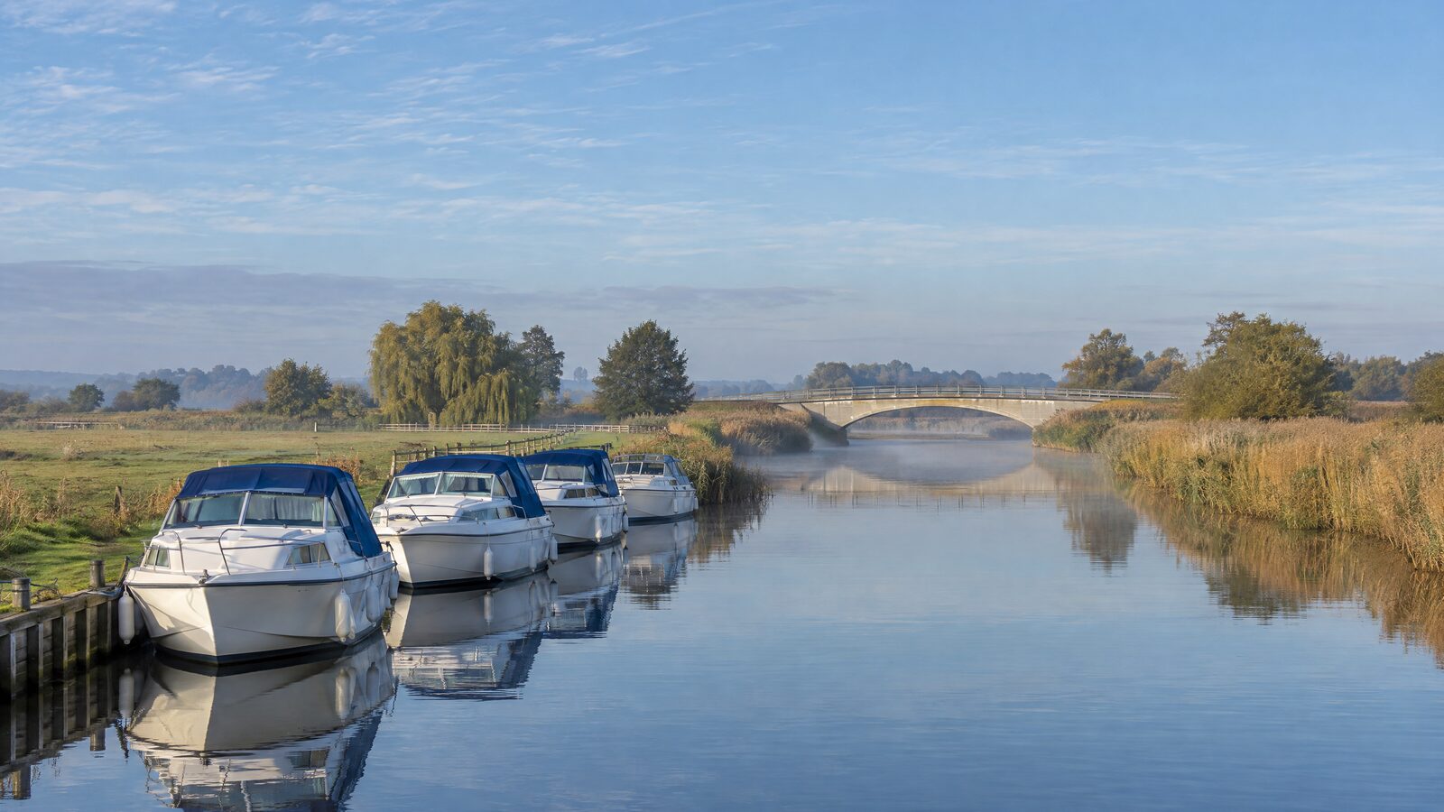 White Broads motor cruisers moored along a reed-fringed river on a misty autumn morning, with a road bridge beyond