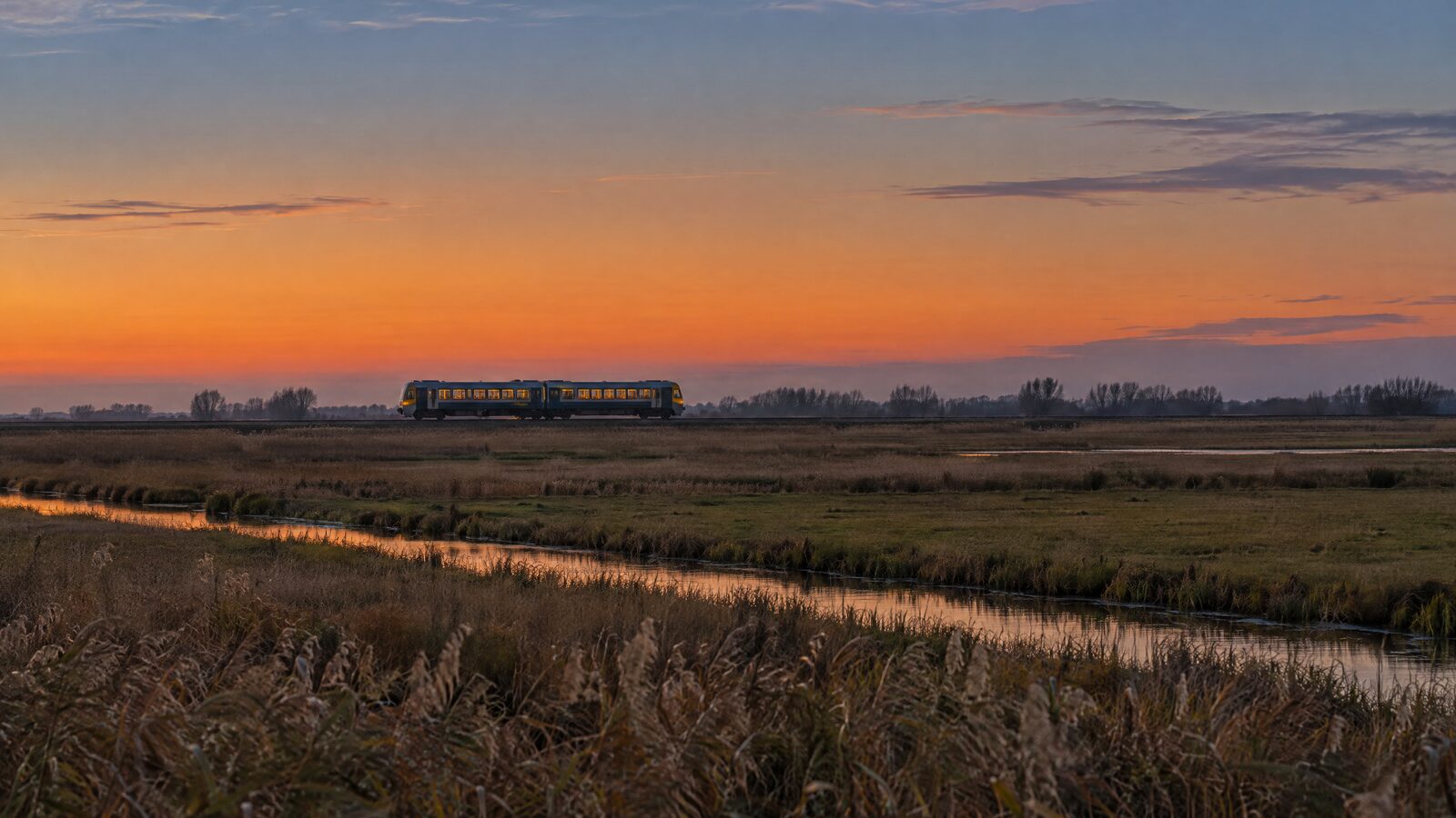 A two-carriage regional train crossing flat Broadland grazing marsh at dusk, with drainage dykes catching the last of the light